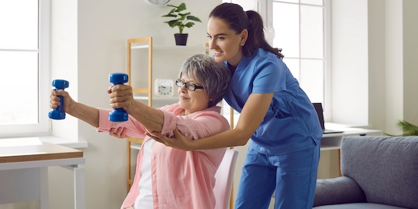 nurse or physiotherapist in scrubs helping a happy retired old woman do fitness exercises with light weight dumbbells at home. 